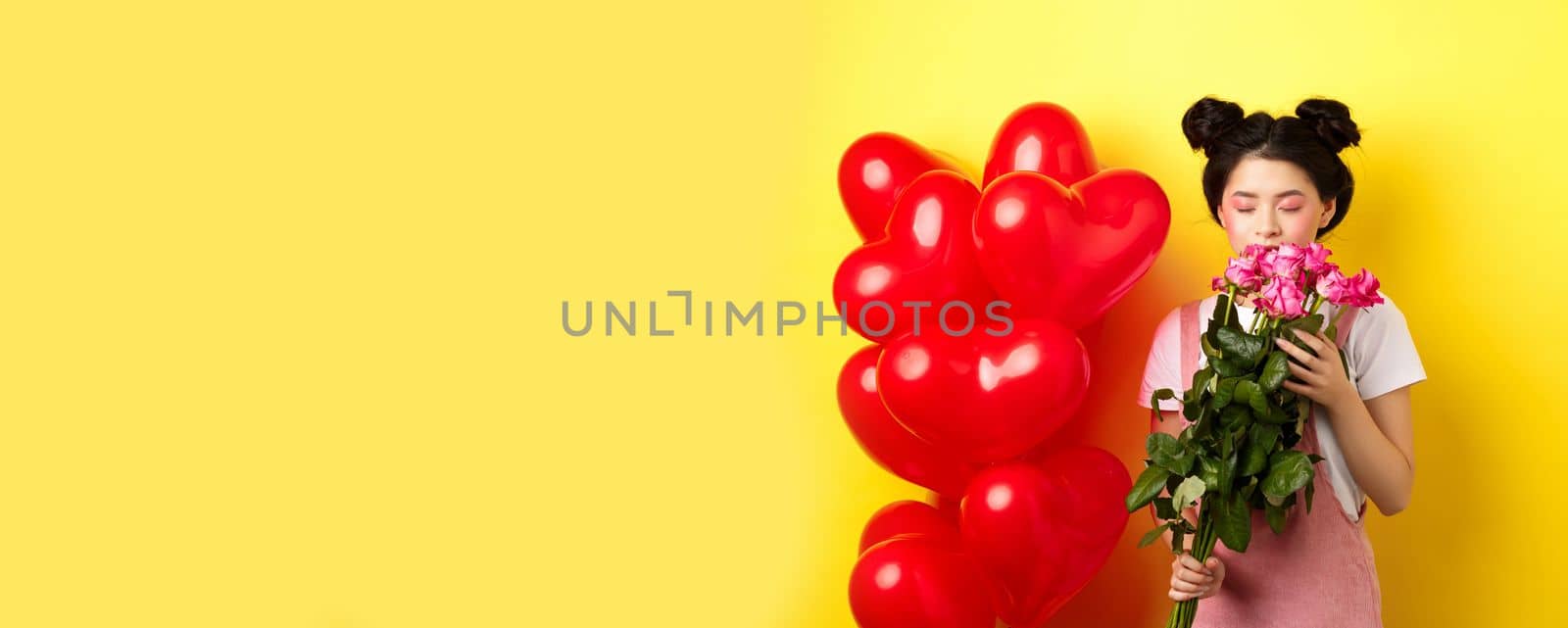 Happy Valentines day. Tender asian girl smelling flower from boyfriend. Girlfriend holding beautiful pink roses, standing near romantic hearts balloons, yellow background.