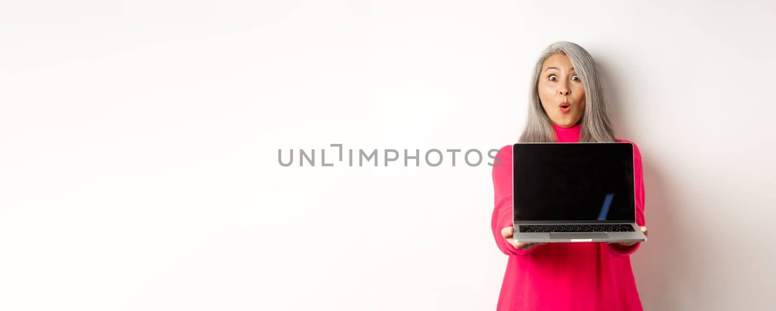 E-commerce concept. Impressed asian senior woman showing blank laptop screen and staring amazed at camera, standing over white background by Benzoix