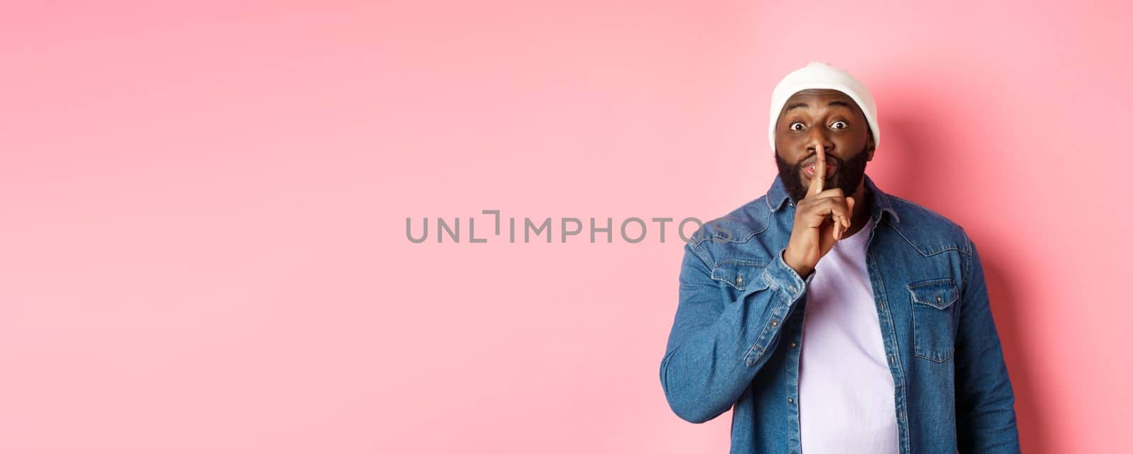 Handsome young black guy in beanie and denim shirt prepare surprise, hushing softly and looking at camera, asking keep quiet, pink background.