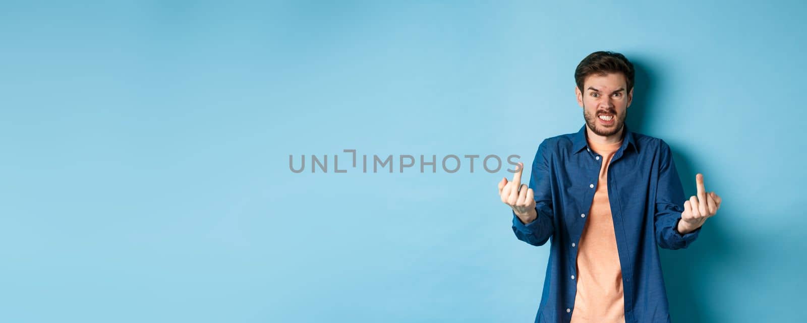 Angry rude guy showing middle finger and saying fuck you, swearing and staring furious at camera, standing on blue background.