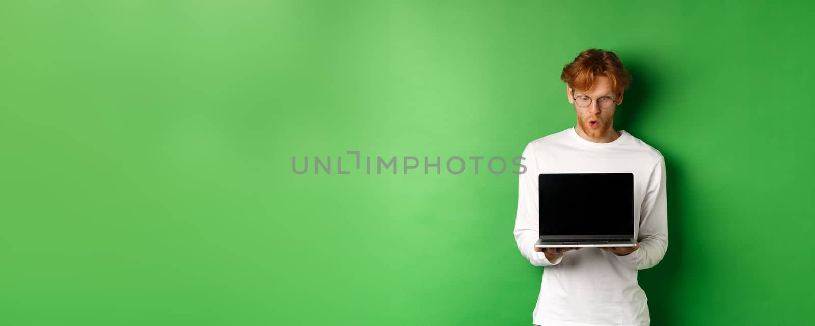 Impressed redhead nerdy guy in glasses showing blank laptop screen and looking at display amazed, standing over green background.