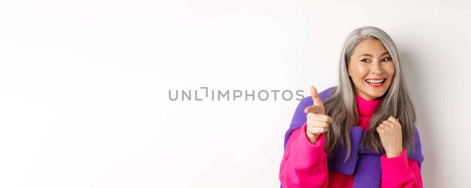 Close-up of happy and cool senior hipster woman, checking out, pointing and looking at person, smiling sideways, standing over white background.