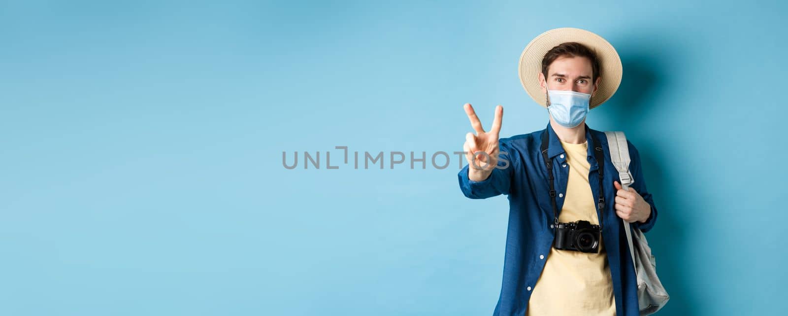 Covid-19, pandemic and travel concept. Positive guy going on vacation, wearing summer hat and medical mask, showing peace sign, standing with backpack and camera on blue background.