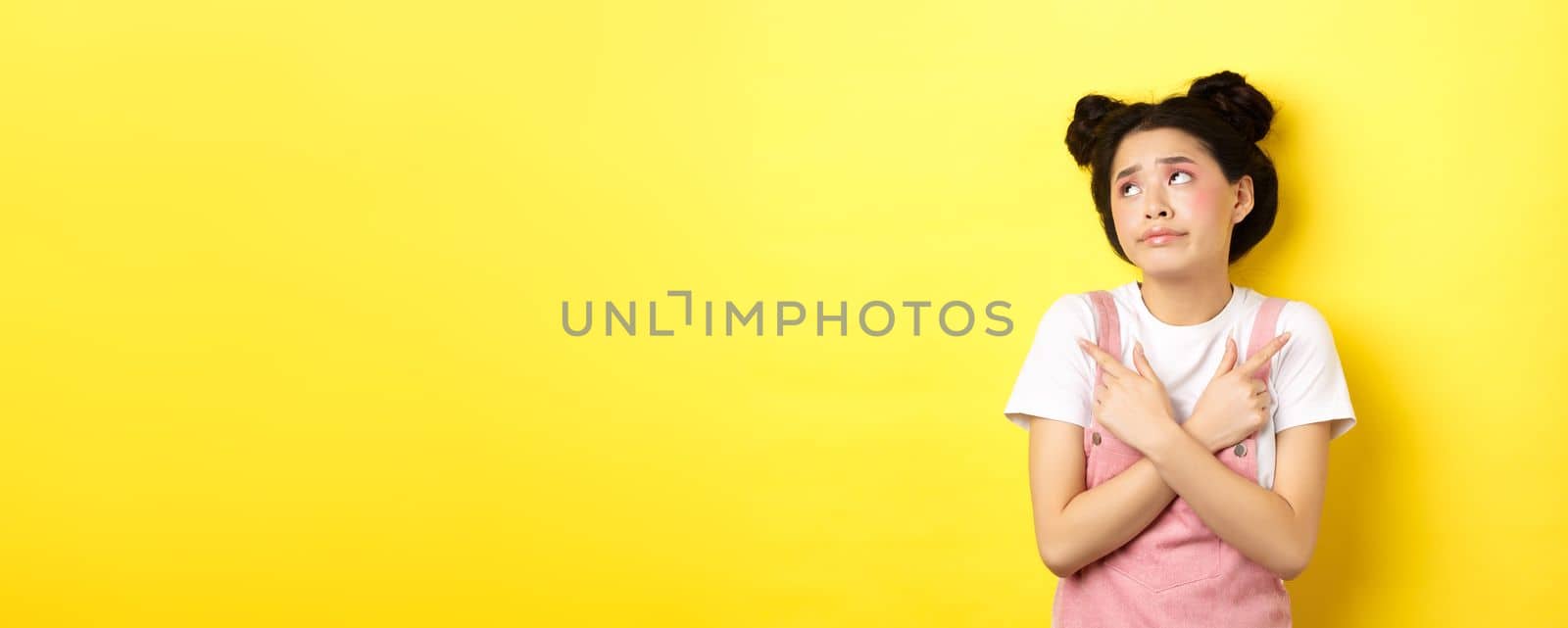 Indecisive asian teen girl troubled to make choice, pointing sideways and looking at logo confused, standing on yellow background.
