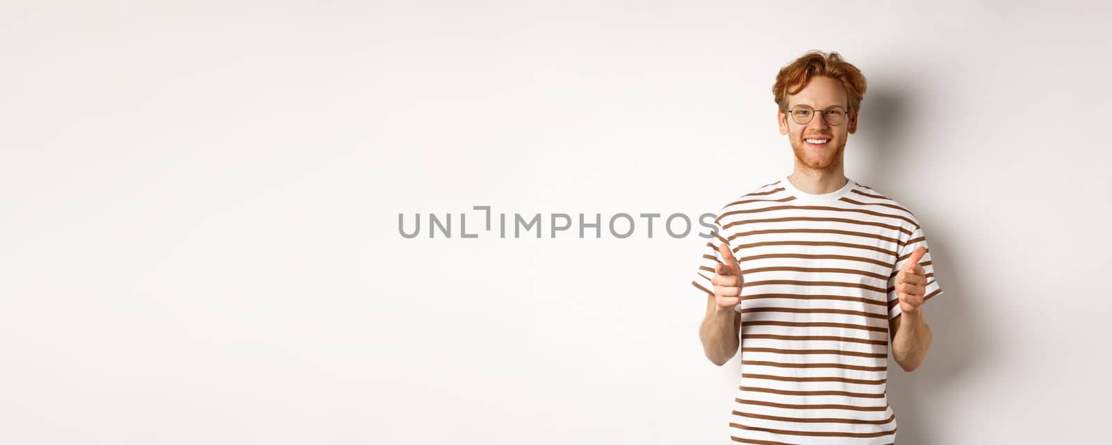 Young man with red hair and glasses pointing fingers at camera and smiling, need you, standing over white background by Benzoix