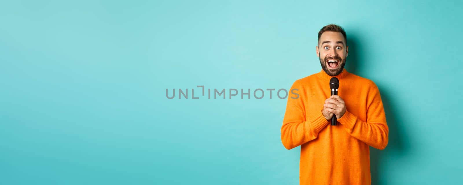 Happy adult man singing karaoke, holding microphone and looking at camera, standing in orange sweater against turquoise background.
