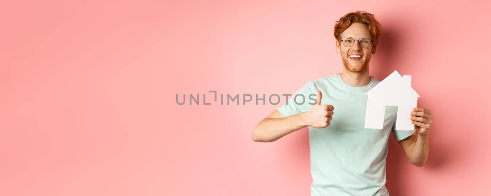 Real estate. Cheerful man in glasses and t-shirt recommending broker agency, showing paper house cutout and thumbs-up, standing over pink background.