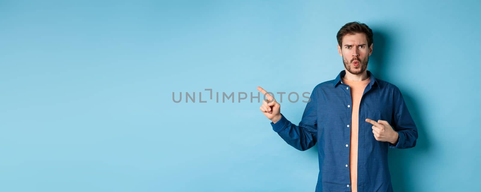 Shocked and offended young man frowning and looking hurt, pointing fingers left at empty space, complaining on something, standing on blue background.