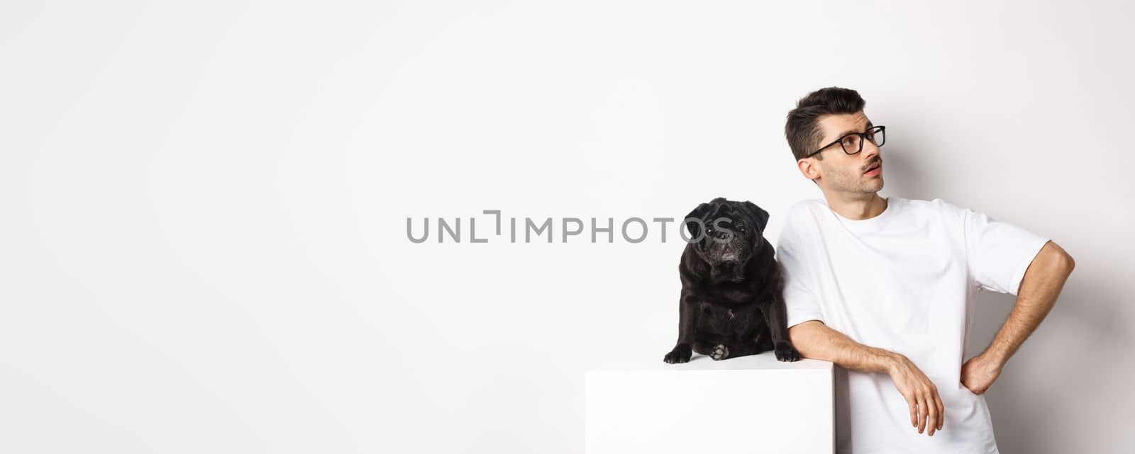 Handsome young man standing near cute black pug, looking right with arrogant expression, standing over white background.