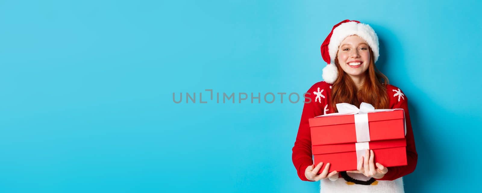 Happy holidays and Christmas concept. Cheerful teen girl with red hair, wearing santa hat and holding xmas presents, by Benzoix