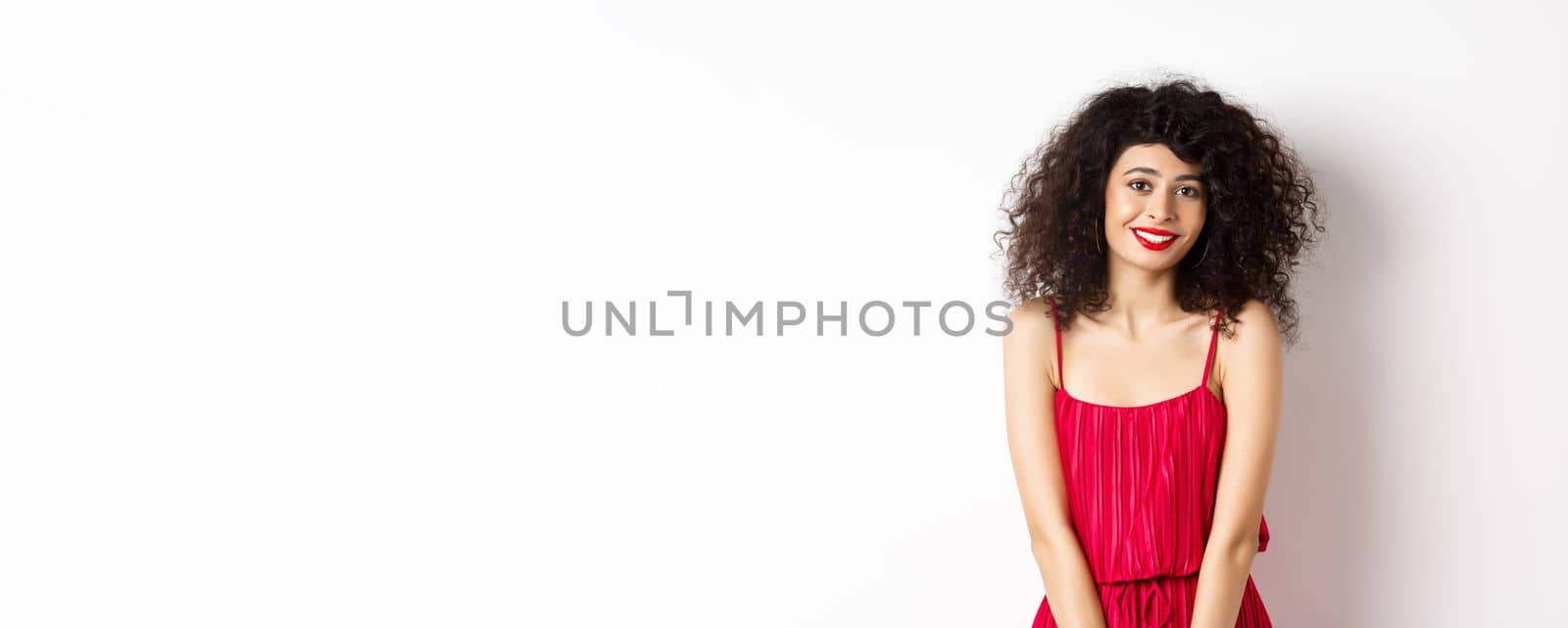 Cute modest lady in red dress and makeup, smiling and blushing, looking at camera, standing over white background.