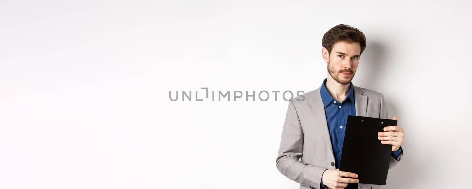 Young male ceo manager holding clipboard with documents and looking at camera, inspecting office, standing on white background.