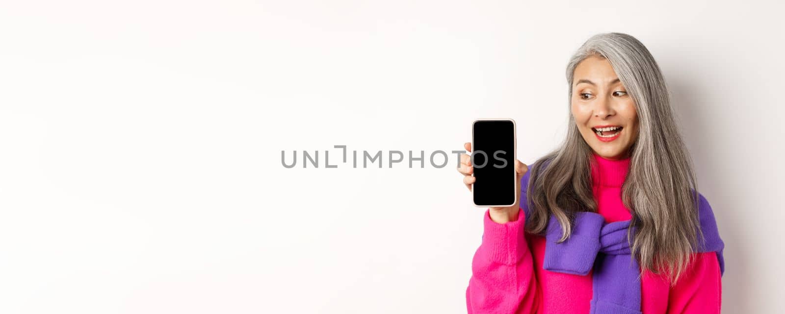 Online shopping. Stylish and beautiful asian senior woman with grey hair, showing blank smartphone screen and looking pleased, standing over white background.