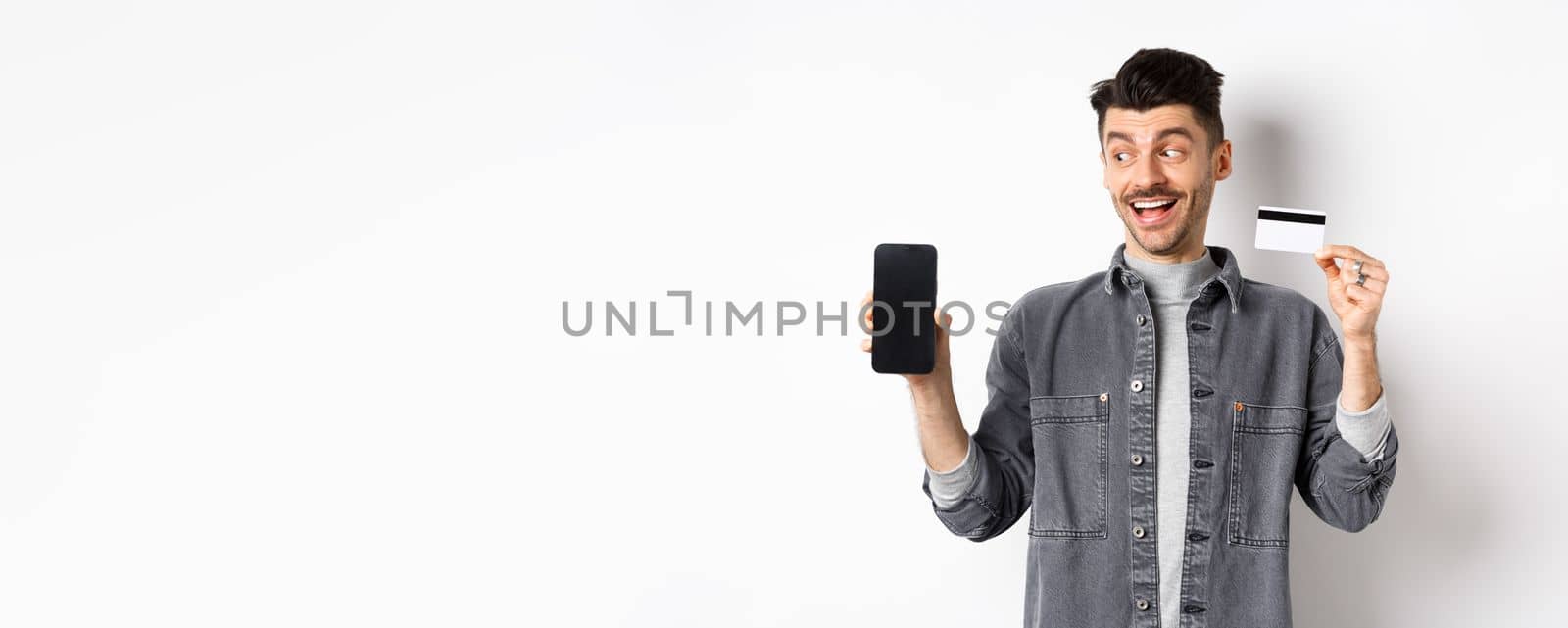 Handsome smiling man in denim jacket looking at empty smartphone screen and showing plastic credit card, good internet offer, standing against white background.