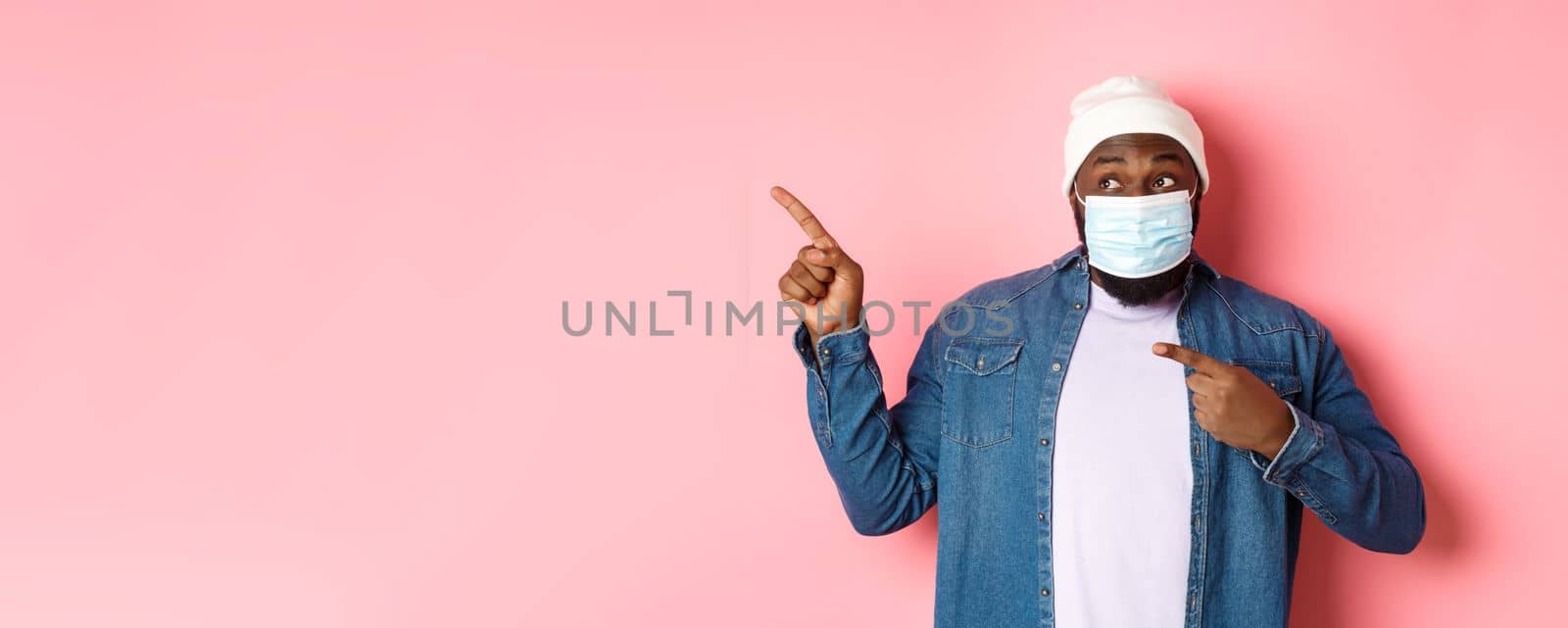 Coronavirus, lifestyle and global pandemic concept. Amused african-american male model in face mask pointing, looking left at promo offer, showing banner, pink background by Benzoix