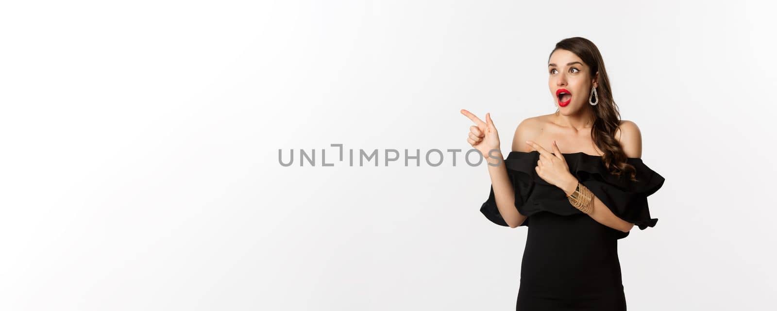 Fashion and beauty. Amazed beautiful woman in black dress open mouth fascinated, pointing fingers and looking left, white background.