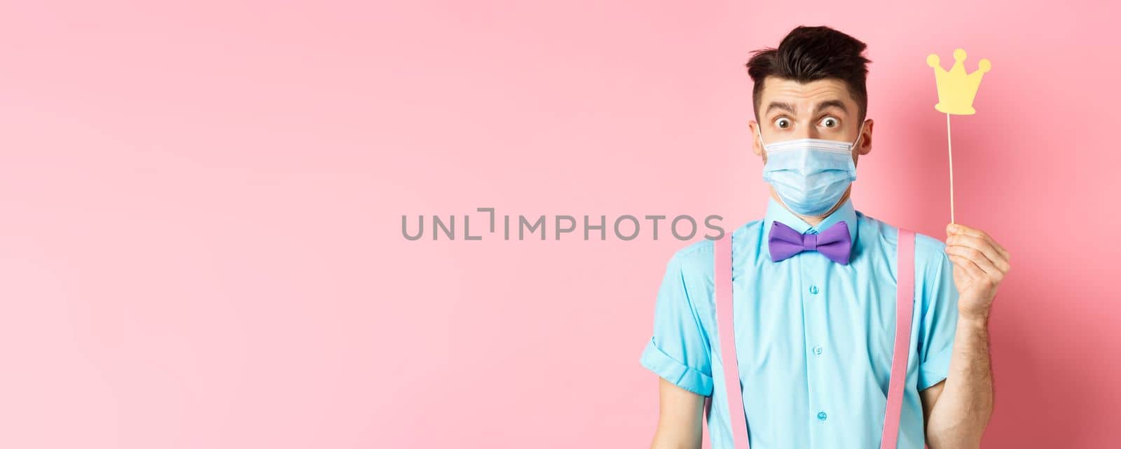 Covid, pandemic and quarantine concept. Cute and silly guy in medical mask looking surprised, holding small crown for party, standing over pink background by Benzoix