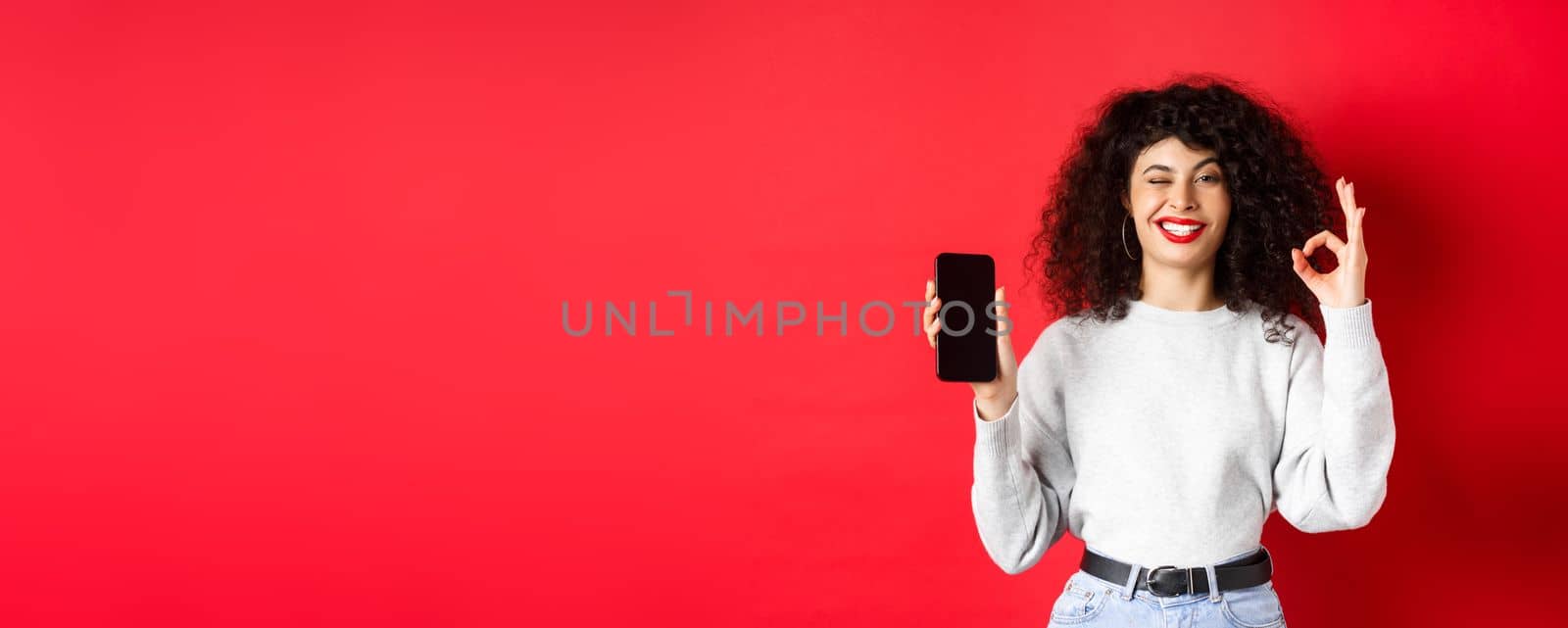 Attractive woman with smartphone, showing okay sign and empty phone sreen, recommending shopping app, standing against red background by Benzoix