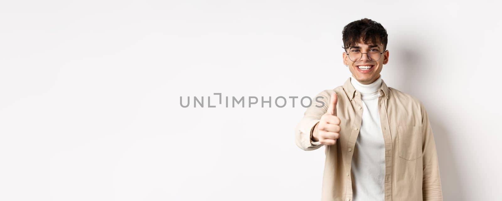 Image of happy young man showing thumb up and smiling satisfied, praise good job, say well done and look pleased, standing on white background.