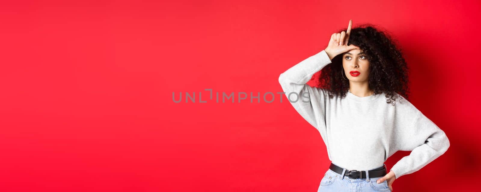 Arrogant woman with curly hair, showing loser sign on forehead and looking aside, mocking someone, standing on red background.