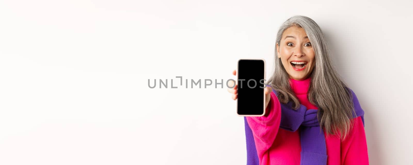 Online shopping. Close up of stylish asian senior woman extending hand with mobile phone, showing blank smartphone screen and smiling, standing over white background by Benzoix
