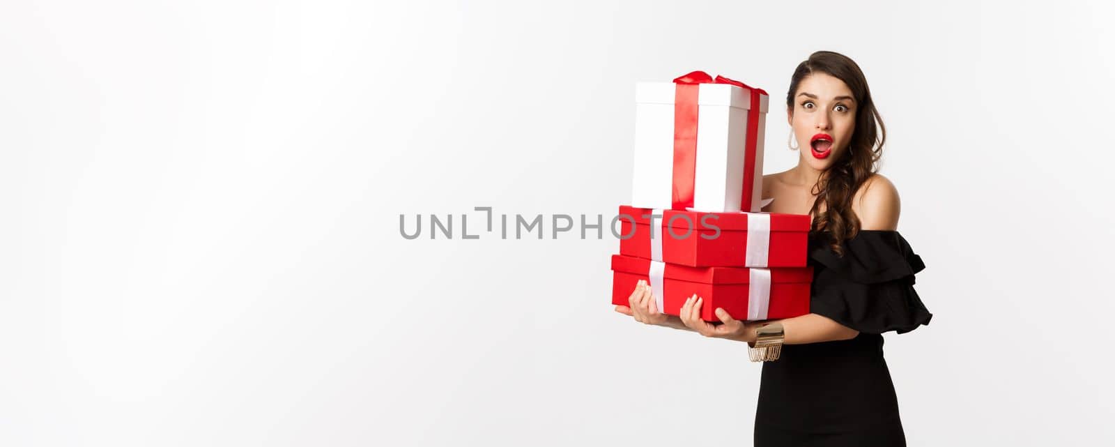 Celebration and christmas holidays concept. Beautiful woman in black dress holding gifts and looking surprised, standing over white background.