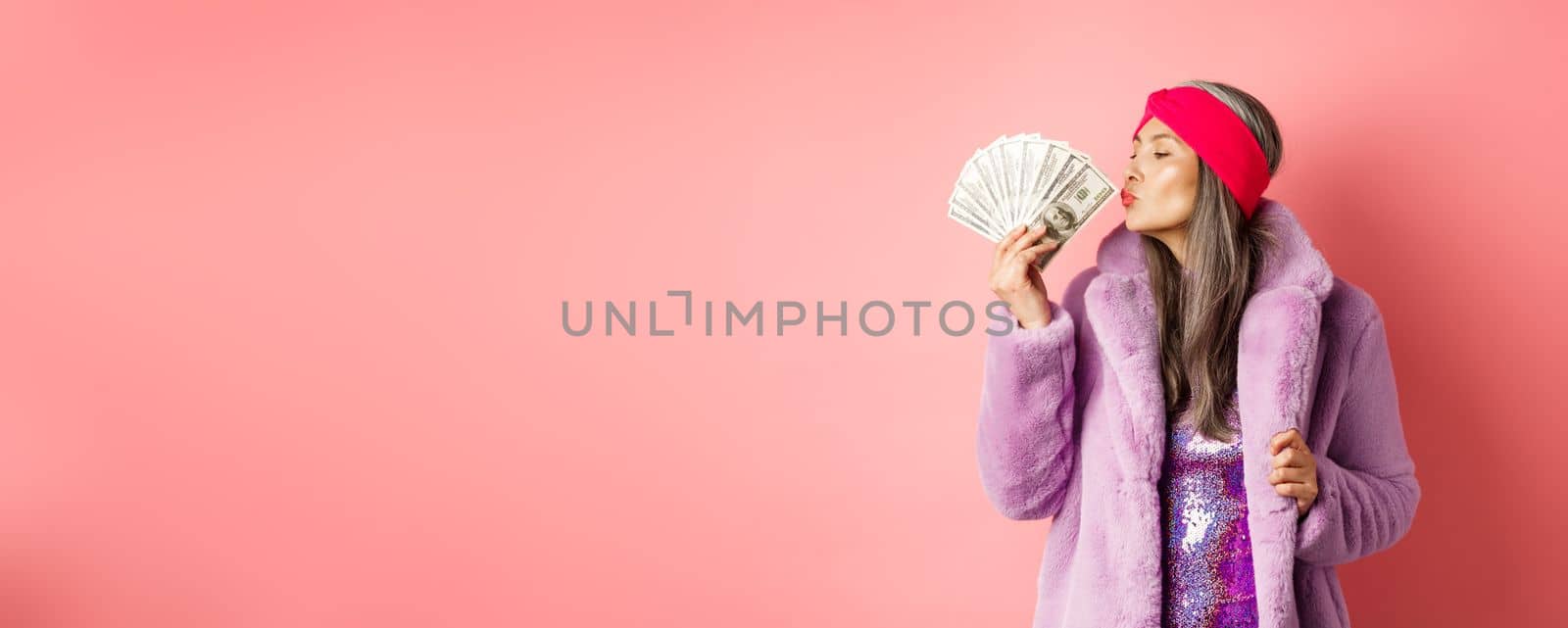 Shopping and fashion concept. Fashionable and rich senior woman kissing dollars money, looking pleased, wearing purple faux fur coat with party dress, pink background.