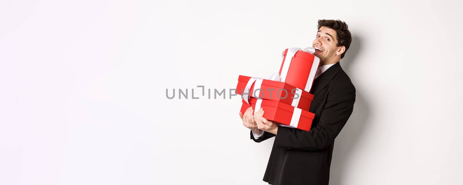 Concept of christmas holidays, celebration and lifestyle. Handsome man in black suit, holding pile of gifts and smiling, wishing happy new year, standing over white background.