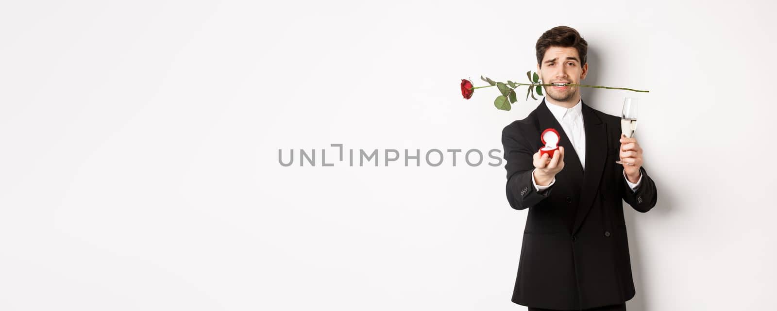 Romantic young man in suit making a proposal, holding rose in teeth and glass of champagne, showing engagement ring, asking to marry him, standing against white background.