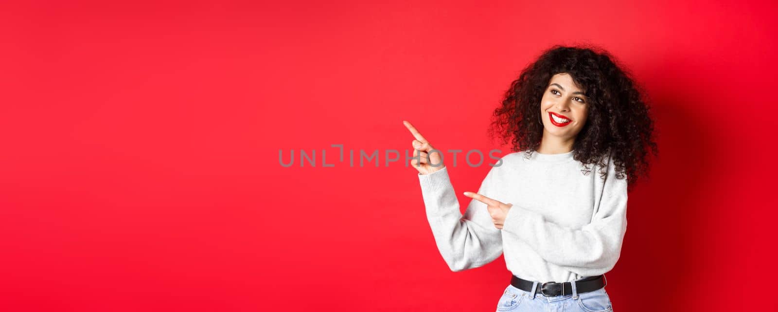 Beautiful european woman with curly hair and makeup, pointing fingers left and looking aside with dreamy smile, checking out promotion deal, red background.