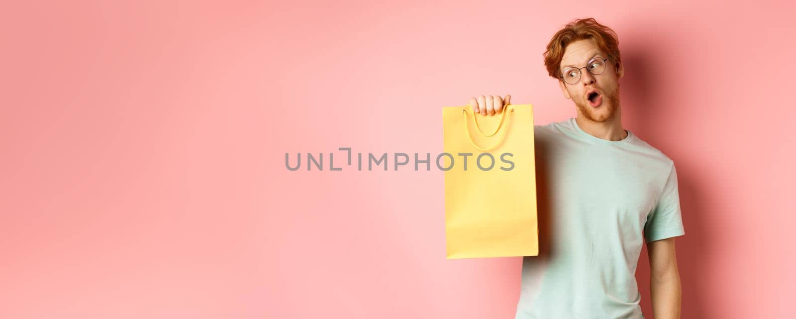 Funny handsome man with red hair, wearing glasses and t-shirt, holding and looking at yellow shopping bag, buying gifts during promo offer, standing over pink background.