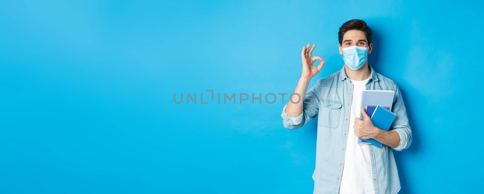 Education, covid-19 and social distancing. Guy student in medical mask looking happy, holding notebooks, showing ok sign, standing over blue background by Benzoix