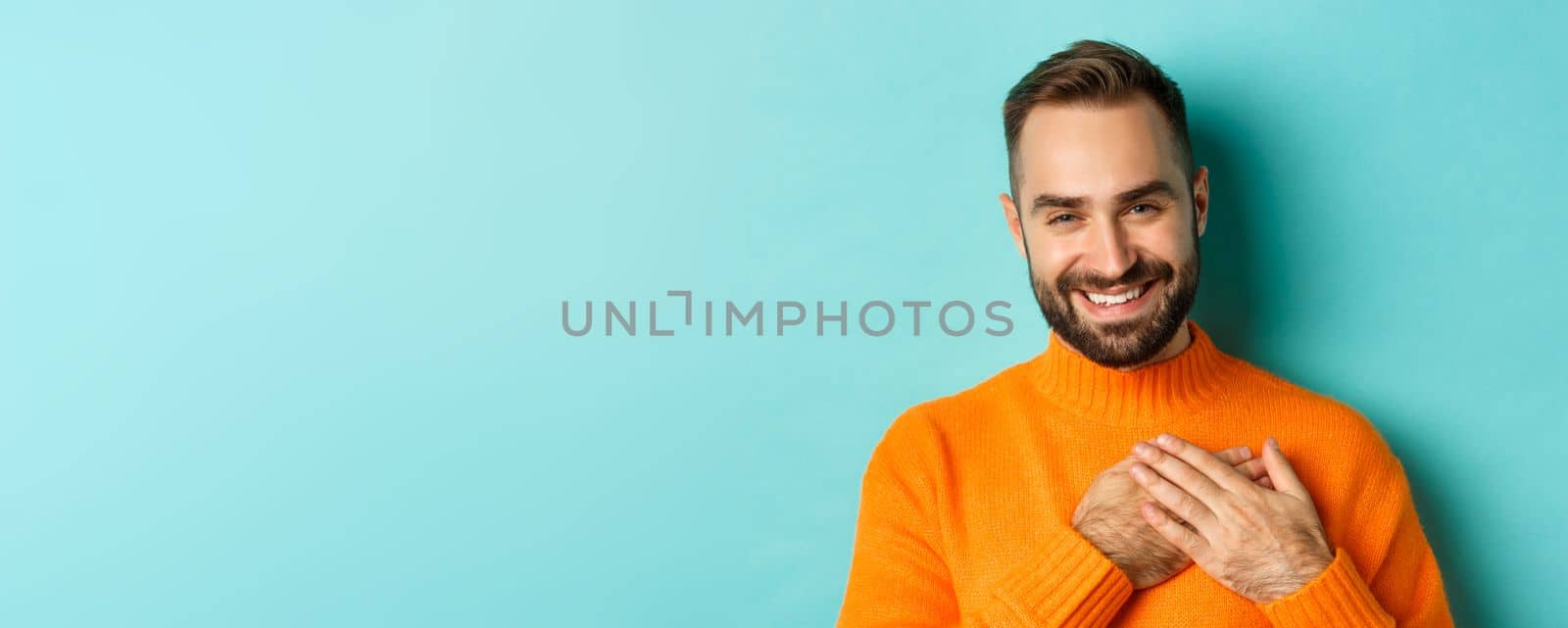 Close-up of handsome young man saying thank you, holding hands on heart and smiling grateful, standing over light blue background.