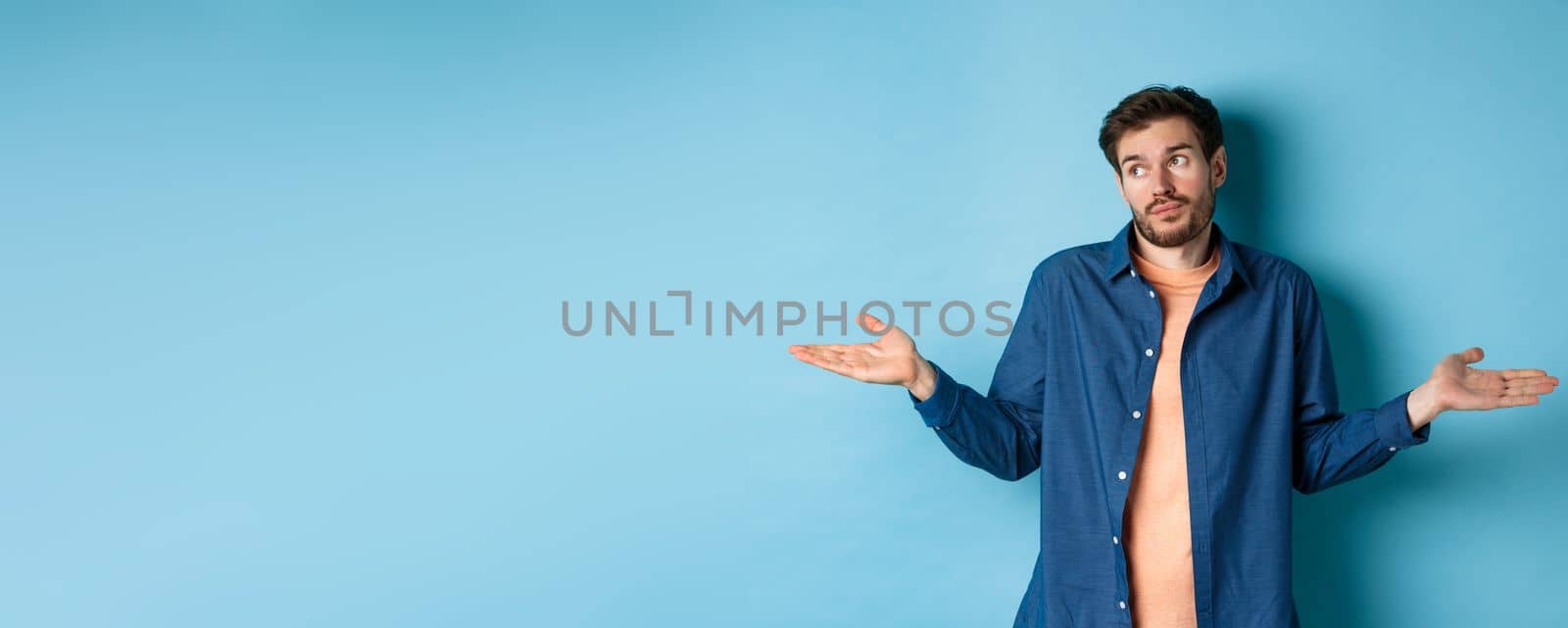 Indecisive young man with beard, look away and shrugging shoulders, know nothing, being clueless, standing on blue background.