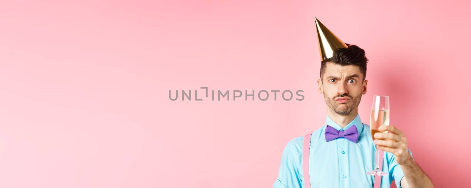 Holidays and celebration concept. Troubled young man in party hat, frowning with doubtful face, raising glass of champagne perplexed, standing on pink background by Benzoix