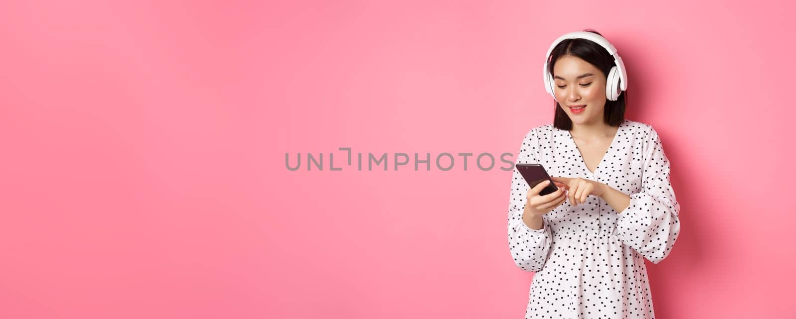 Beautiful asian woman texting message on smartphone, listening music in headphones, standing over pink background.
