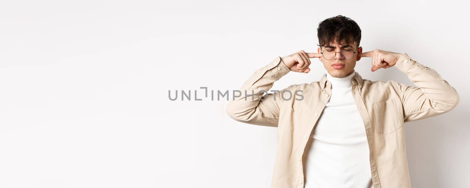 Annoyed young man shut his ears, block annoying sound, unwilling to listen, standing on white background.