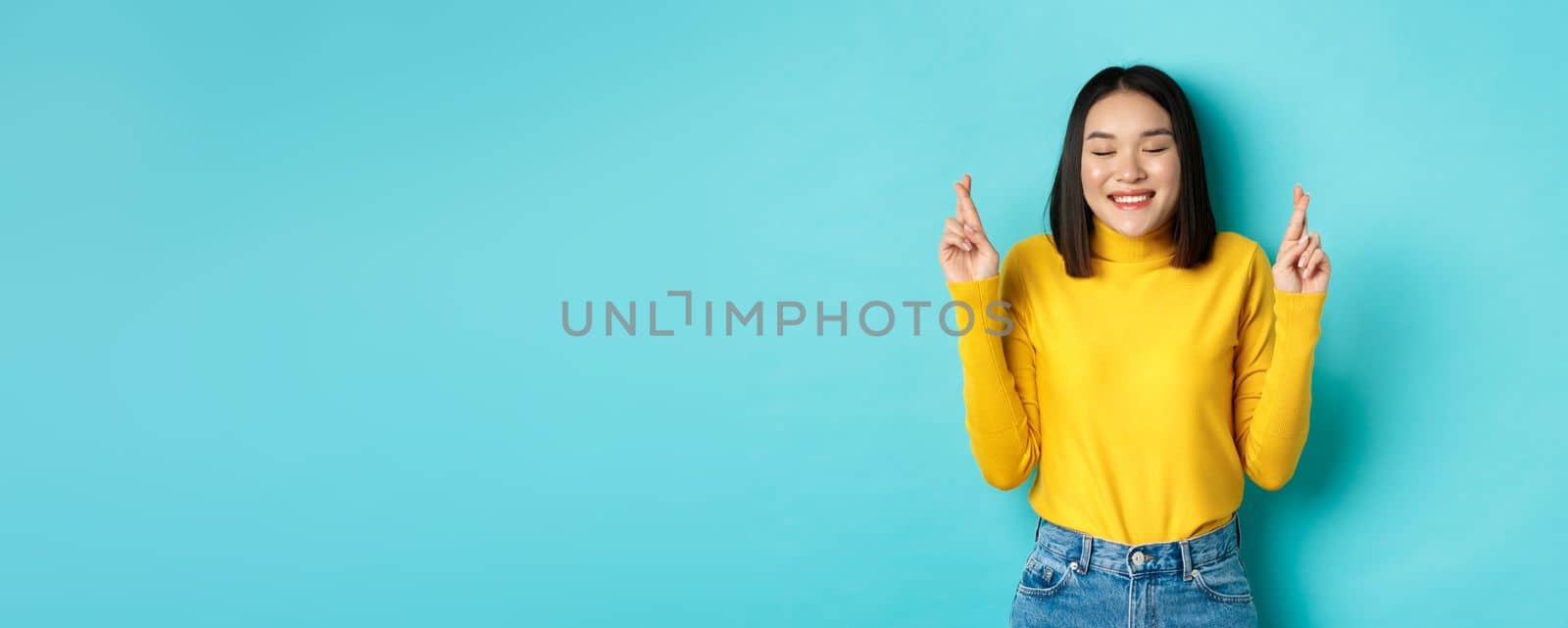 Hopeful asian woman dreaming, cross fingers for good luck and making wish, praying or supplicating, smiling with closed eyes, standing over blue background.