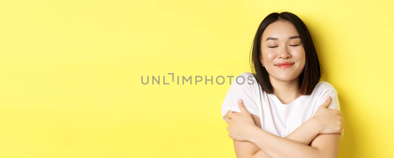 Close up of romantic asian girl hugging herself and dreaming, close eyes and smile while imaging something tender, standing over yellow background by Benzoix