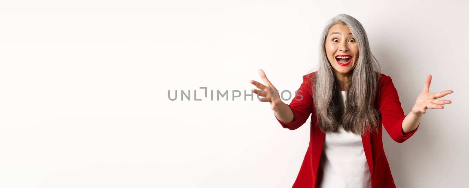 Surprised and friendly asian woman reaching hands forward and smiling, welcoming you, greeting someone, standing over white background.