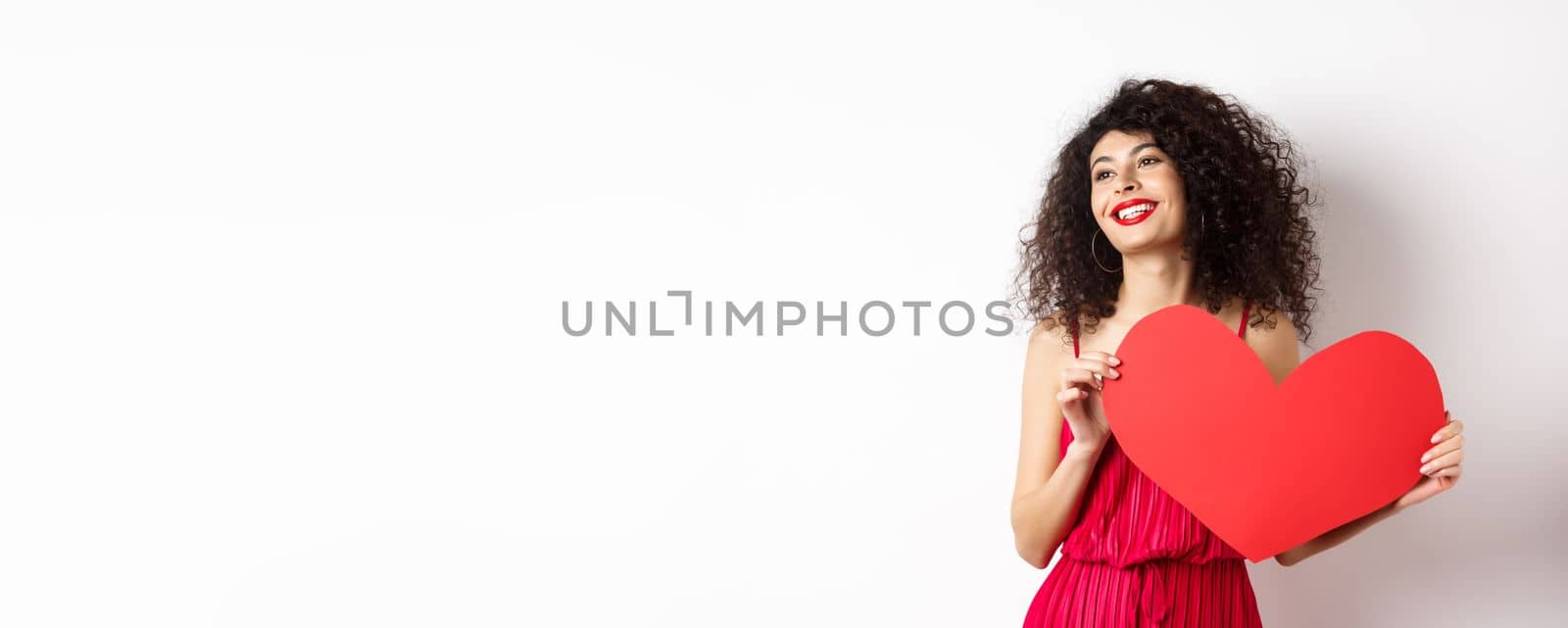 Romantic woman in dress and makeup, looking aside with dreamy face and love, showing big red valentines heart card, standing on white background.