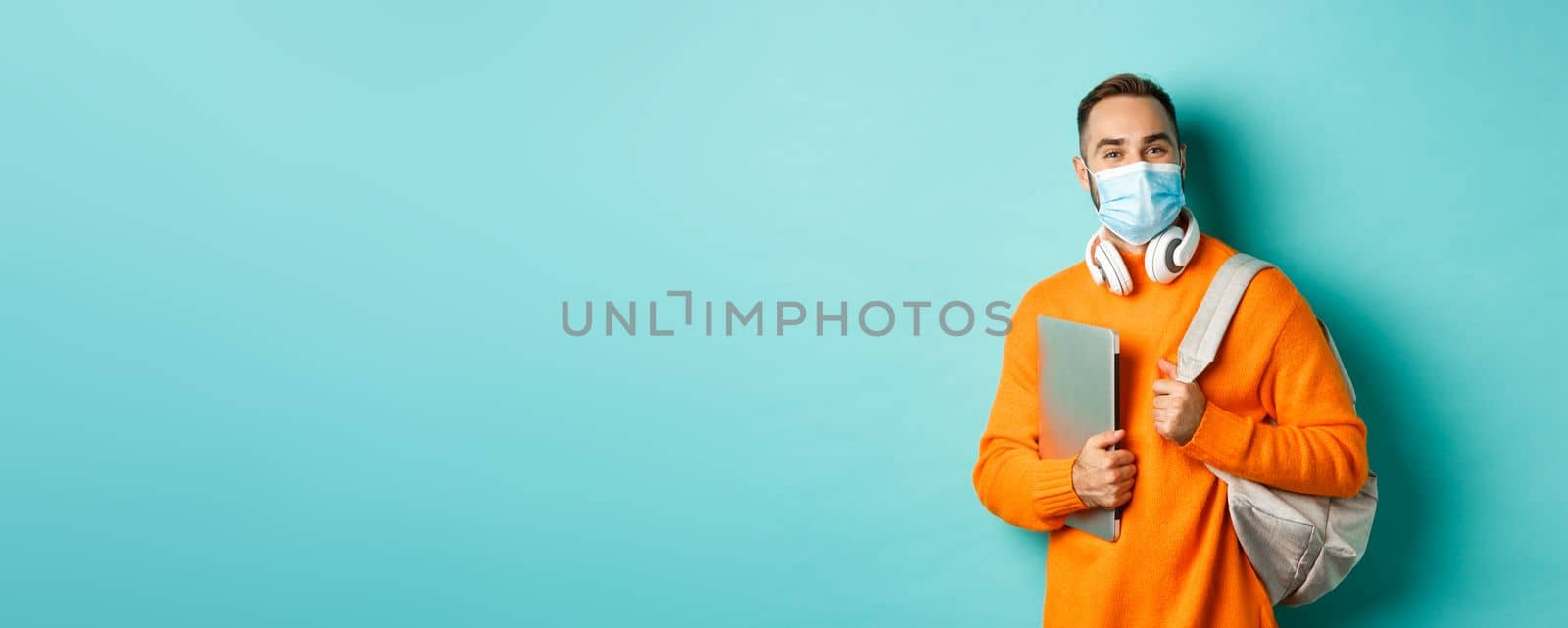 Handsome student in face mask, holding backpack and laptop, going work, standing over light blue background by Benzoix