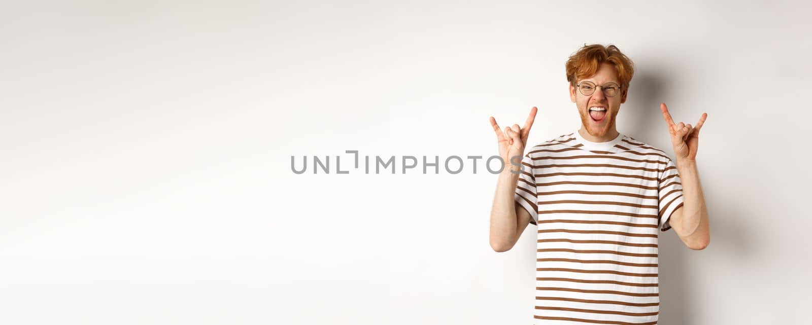 Funny and happy redhead man having fun, showing rock-n-roll horn and sticking tongue, enjoying party, standing over white background by Benzoix