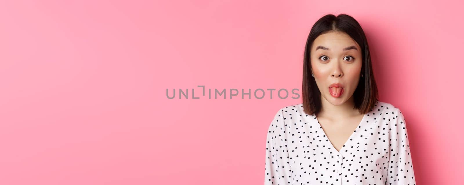 Beauty and lifestyle concept. Close-up of funny and cute asian woman showing tongue, staring at camera silly, standing over pink background by Benzoix