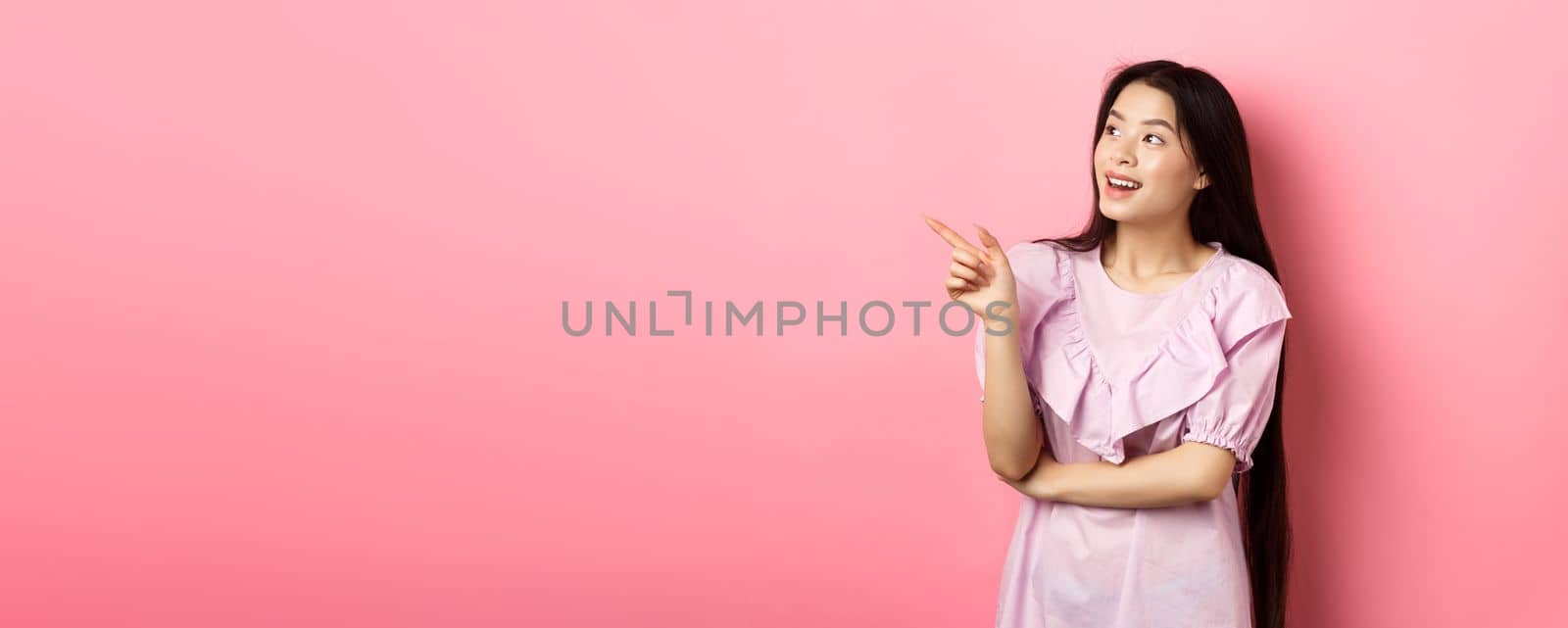 Excited smiling asian woman in dress, pointing and looking left at logo, checking out promotion, standing on pink background.