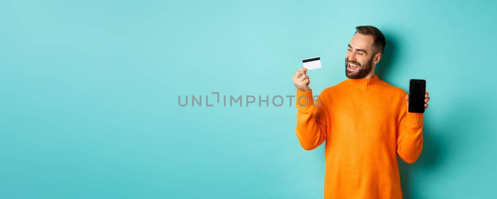 Online shopping. Satisfied man using credit card and showing mobile screen, looking pleased, standing over light blue background by Benzoix