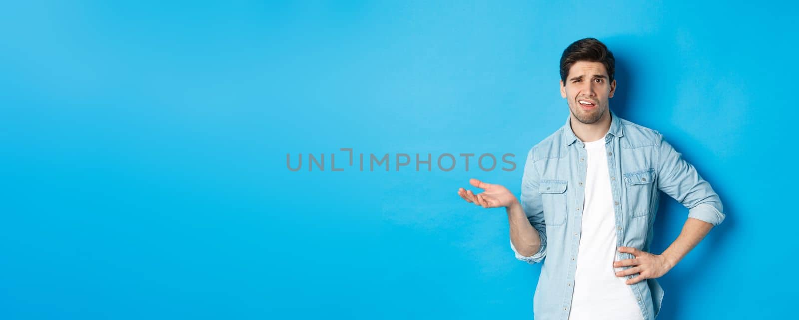 Careless man looking at something disappointing, complaining and judging bad product, standing unamused against blue background.