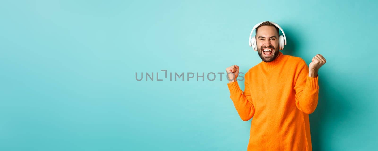 Happy adult man in orange sweater, looking up and listening music in headphones, standing over blue background.