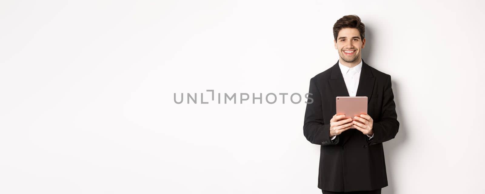 Image of handsome businessman in trendy suit, holding digital tablet and smiling, standing against white background.