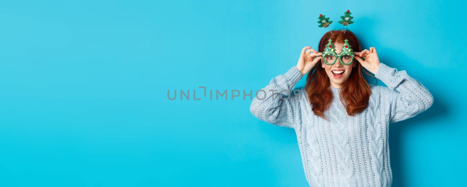Winter holidays and Christmas sales concept. Beautiful redhead female model celebrating New Year, wearing funny party headband and glasses, smiling silly, blue background.